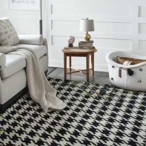 Black and off-white houndstooth rug on gray flooring beside a white armchair and a wooden table.