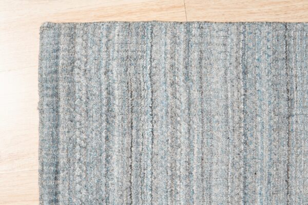 The edge of a textured gray and light blue striped rug lies on pale blonde flooring.