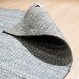 Textured gray and blue striped rug is curled back, exposing its underside on light wood flooring.