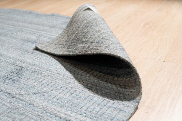 Textured gray and blue striped rug is curled back, exposing its underside on light wood flooring.
