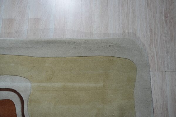 A soft pile rug displays abstract curves in beige, gold, and brown tones on light wood plank flooring.