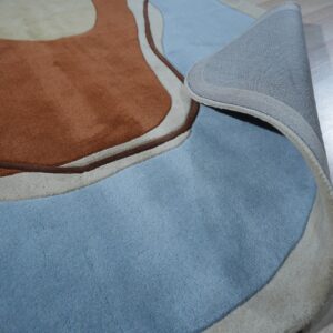 Abstract brown and blue high-pile rug partially rolled back to show the gray fabric backing on light wood flooring.