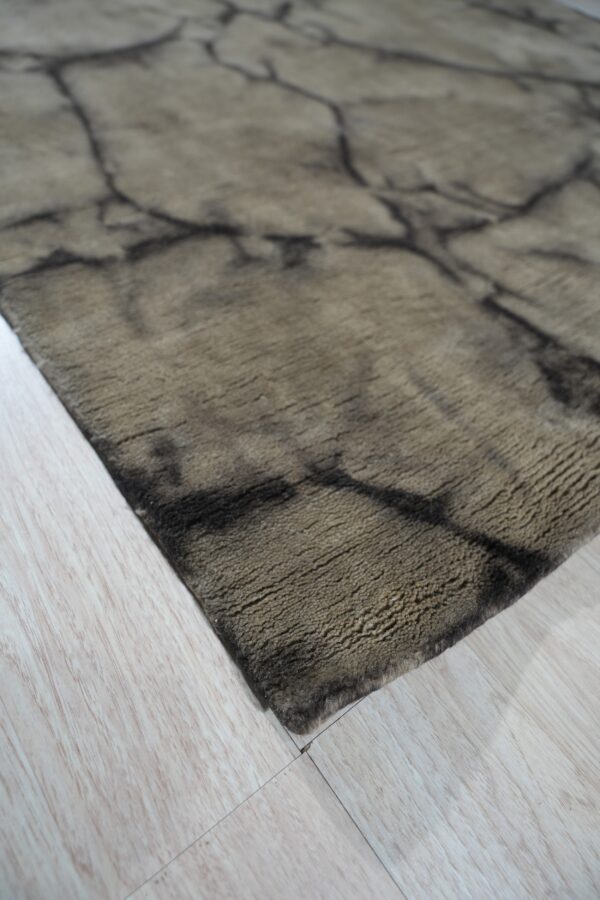 Textured khaki rug with a dark, cracked, abstract pattern rests on light wood-look flooring.