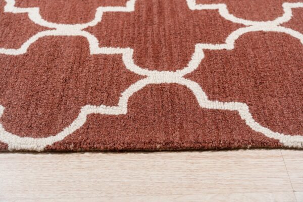 A close-up of a rust rug with a cream geometric trellis pattern on light wood flooring.