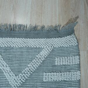 Light gray raised geometric patterns overlay a flat woven gray rug with fringe on wood planks.