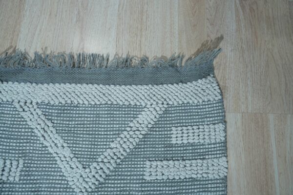 Light gray raised geometric patterns overlay a flat woven gray rug with fringe on wood planks.