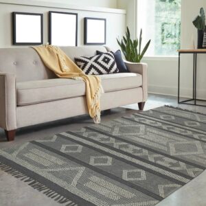 Geometric charcoal and gray flatweave rug with fringe is styled beneath a light beige sofa on concrete flooring.