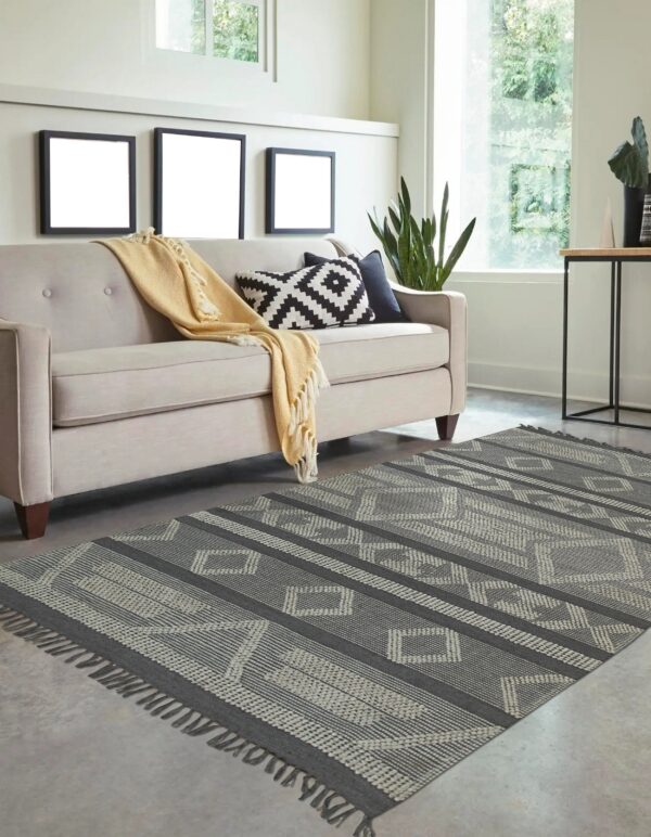 Geometric charcoal and gray flatweave rug with fringe is styled beneath a light beige sofa on concrete flooring.
