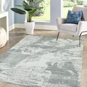 Distressed gray and white abstract rug sits on chevron wood flooring between a large plant and a light gray armchair.