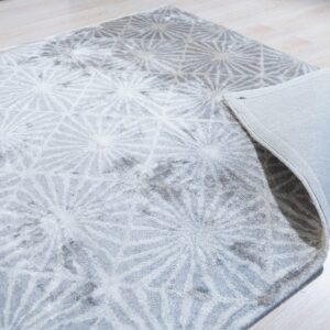 High-pile gray rug featuring white radial patterns rests on light wood flooring, with one corner lifted.
