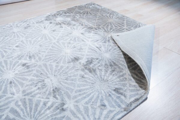 High-pile gray rug featuring white radial patterns rests on light wood flooring, with one corner lifted.