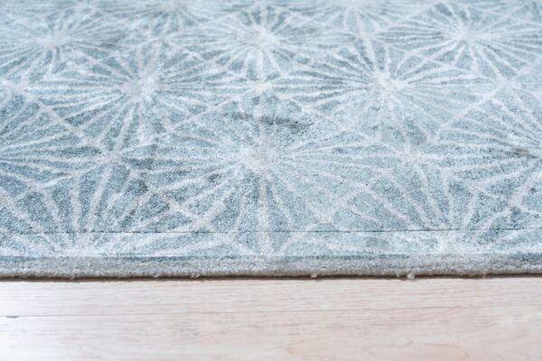 Textured, soft blue rug with a repeated white radial pattern rests on light blonde wood flooring.