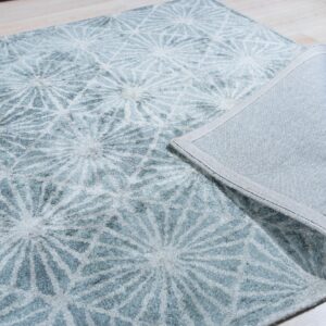 Textured light blue rug with geometric white patterns folded to reveal the gray fabric backing on light wood flooring.