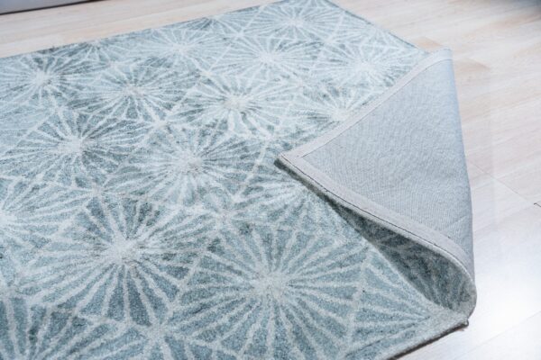 Textured light blue rug with geometric white patterns folded to reveal the gray fabric backing on light wood flooring.