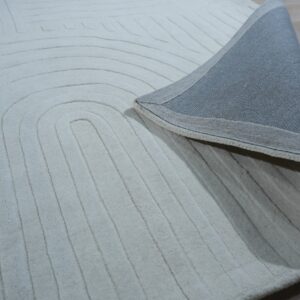 Cream-colored rug with carved concentric lines is folded back, showing the woven gray backing on a light wood floor.