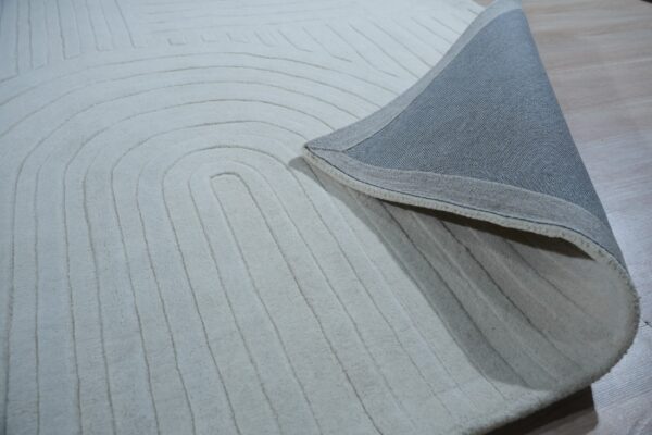 Cream-colored rug with carved concentric lines is folded back, showing the woven gray backing on a light wood floor.