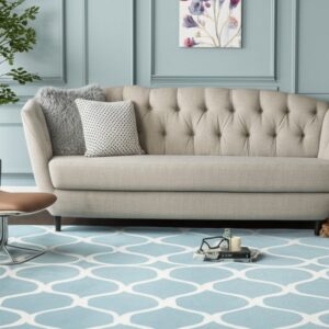 A blue and white geometric patterned rug lies beneath a tufted beige sofa and light blue paneled walls.