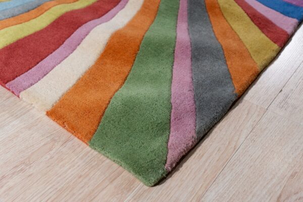 A low-pile rug featuring wide, wavy, multi-colored stripes rests diagonally across light wood flooring.
