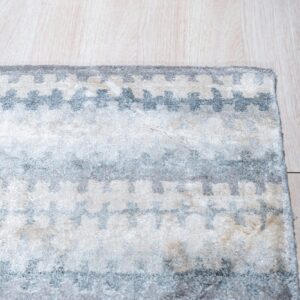 Corner of a soft pile rug displaying repeating light gray and ivory geometric shapes on blonde wood floor.
