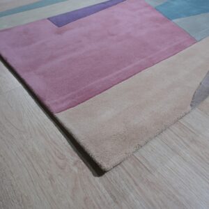 Modern low-pile rug with geometric blocks in pink, beige, blue, and purple on light wood flooring.