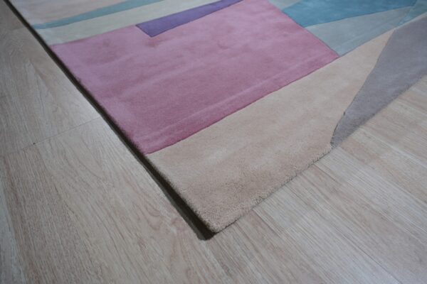 Modern low-pile rug with geometric blocks in pink, beige, blue, and purple on light wood flooring.