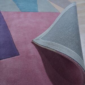 Geometric rug in pink, purple, blue, and beige colors, with the woven backing visible on light wood flooring.