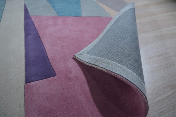 Geometric rug in pink, purple, blue, and beige colors, with the woven backing visible on light wood flooring.