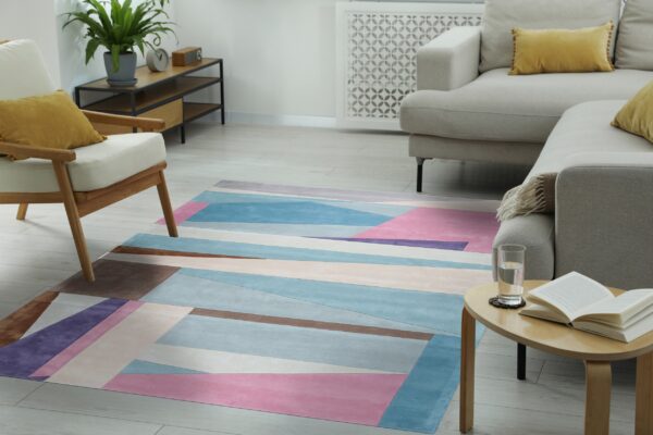 Geometric rug in blue, pink, purple, and brown lies on light flooring beneath a beige sofa and wooden chair.