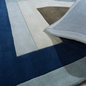 Abstract geometric rug in deep blue, beige, and gray, with the woven backing visible on light wood flooring.