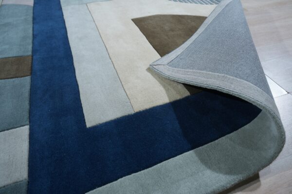 Abstract geometric rug in deep blue, beige, and gray, with the woven backing visible on light wood flooring.