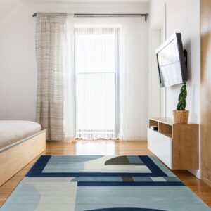 Abstract rug featuring navy, blue, cream, and brown shapes rests on the wood floor of a bright bedroom.