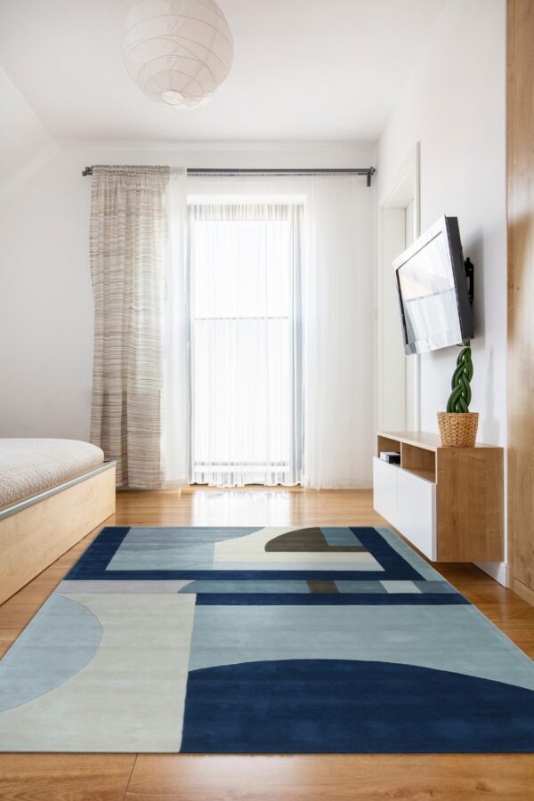 Abstract rug featuring navy, blue, cream, and brown shapes rests on the wood floor of a bright bedroom.