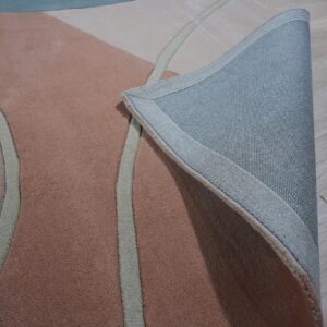 Abstract rug in pink, beige, and blue is folded back, showing its woven gray backing on wood floor.