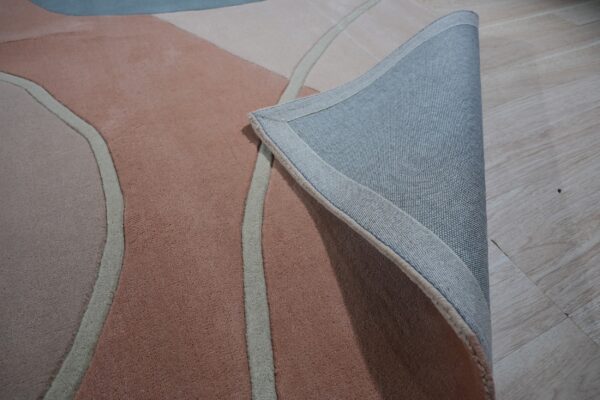 Abstract rug in pink, beige, and blue is folded back, showing its woven gray backing on wood floor.