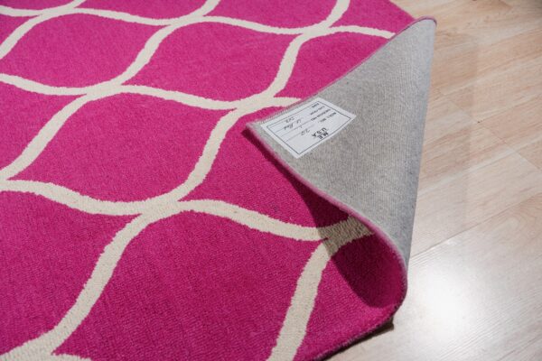 A curled magenta and off-white geometric pattern rug with a tag, exposing the backing on light wood flooring.