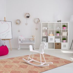 A peach and cream flatwoven rug with a geometric diamond pattern is centered in a bright white nursery.