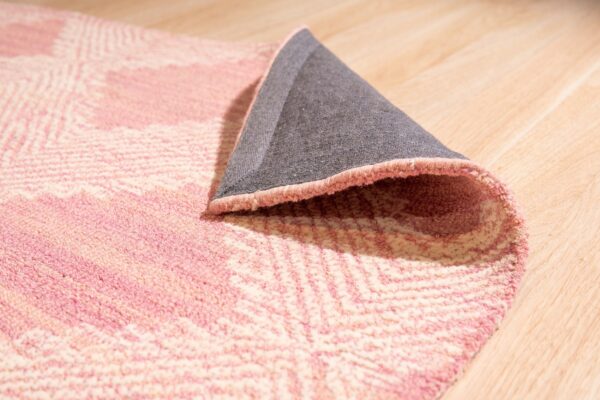 A pink and cream patterned rug with a medium pile is rolled back on light wood flooring, revealing a gray backing.