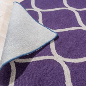 A purple rug with a light gray trellis pattern is peeled back, revealing its woven backing on light wood flooring.