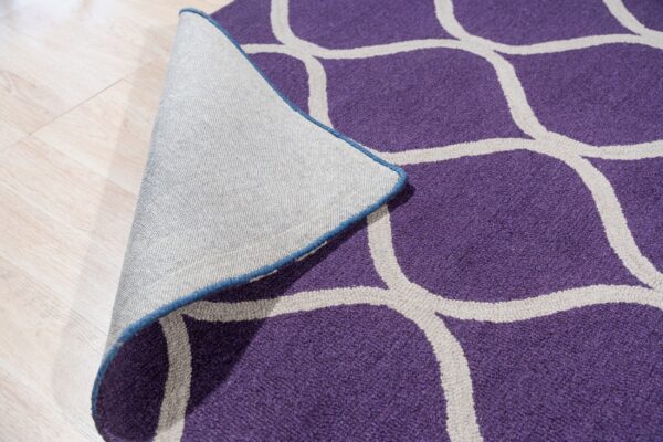 A purple rug with a light gray trellis pattern is peeled back, revealing its woven backing on light wood flooring.