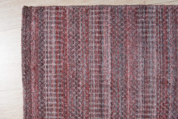 A corner view of a woven red and grey heathered rug with subtle white grid lines on light wood flooring.