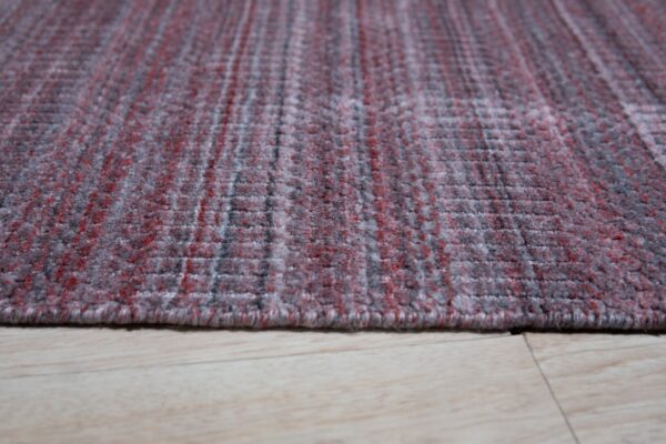 Textured low-angle view of a red and gray heathered rug edge resting on light wood plank flooring.