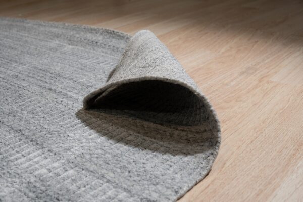 Mottled gray textured rug rolled back on light wood flooring shows the pile and backing.