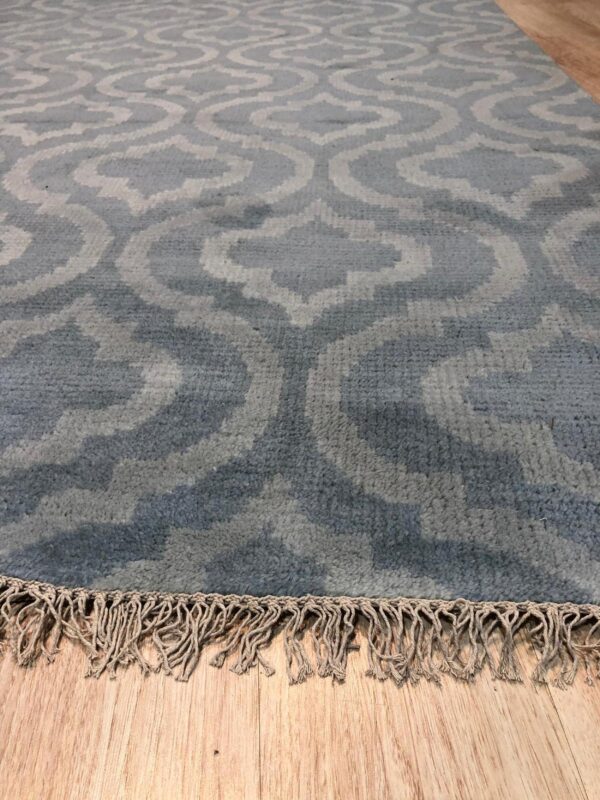 Angled view of a blue and gray trellis rug with natural braided fringe on wood flooring.