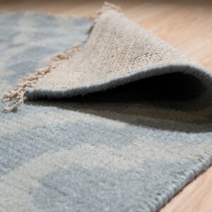 Close-up of a low pile blue and light gray patterned rug folded back on light wood flooring.