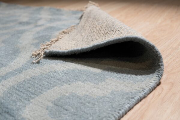 Close-up of a low pile blue and light gray patterned rug folded back on light wood flooring.