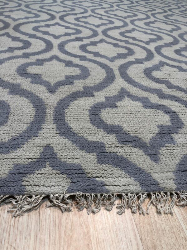 Textured light gray rug with a dark blue interlocking geometric pattern and fringe resting on wood flooring.