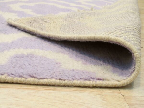 Close-up of a lavender and cream thick pile rug rolled back, showing its woven backing and binding on wood flooring.