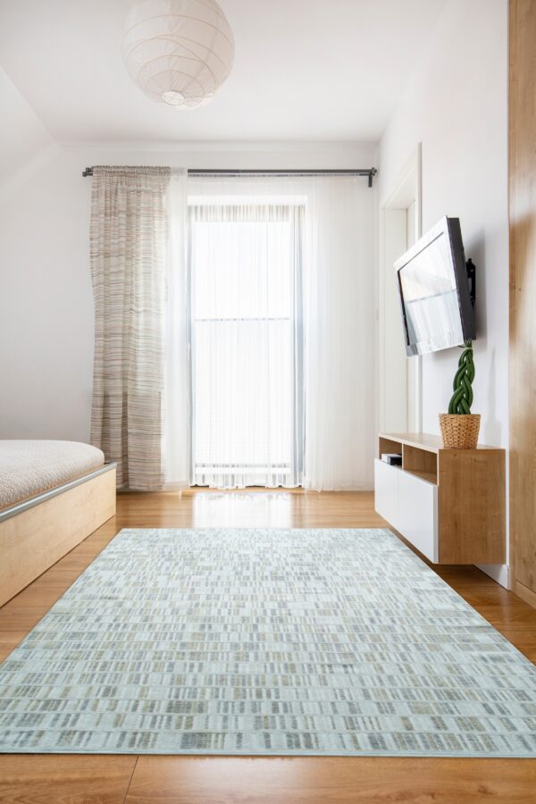 Light blue rug with a geometric dash pattern sits on warm wood floors in a sunny, white bedroom.