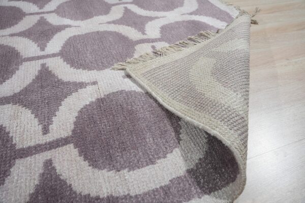Mauve and cream geometric rug, showing woven backing and fringe, rests on light wood flooring.