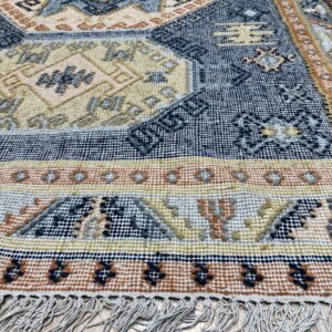 Geometric flatweave rug in blue, gold, and terracotta with white fringe on a wooden floor.